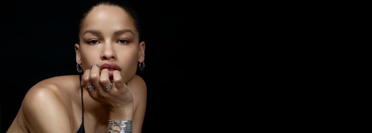 Woman with hand on chin against a black background wearing silver bracelets and rings